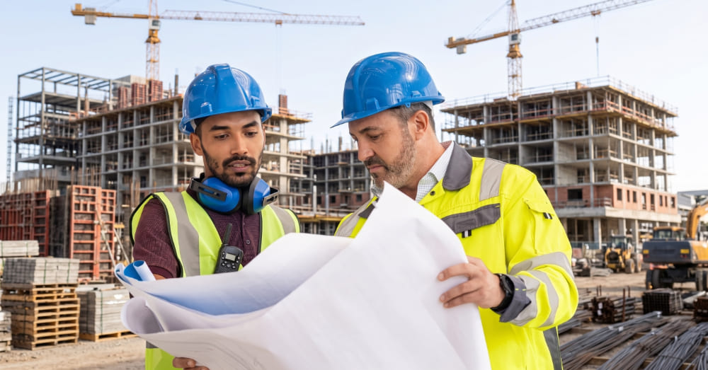 Two construction workers in blue hard hats and safety vests looking at blueprints on a job site, representing employees who use an hours bank arrangement for continuous health insurance.